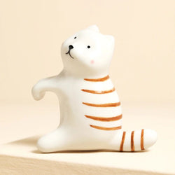 A small white cat figurine with orange stripes, sitting on a beige surface against a warm beige background.