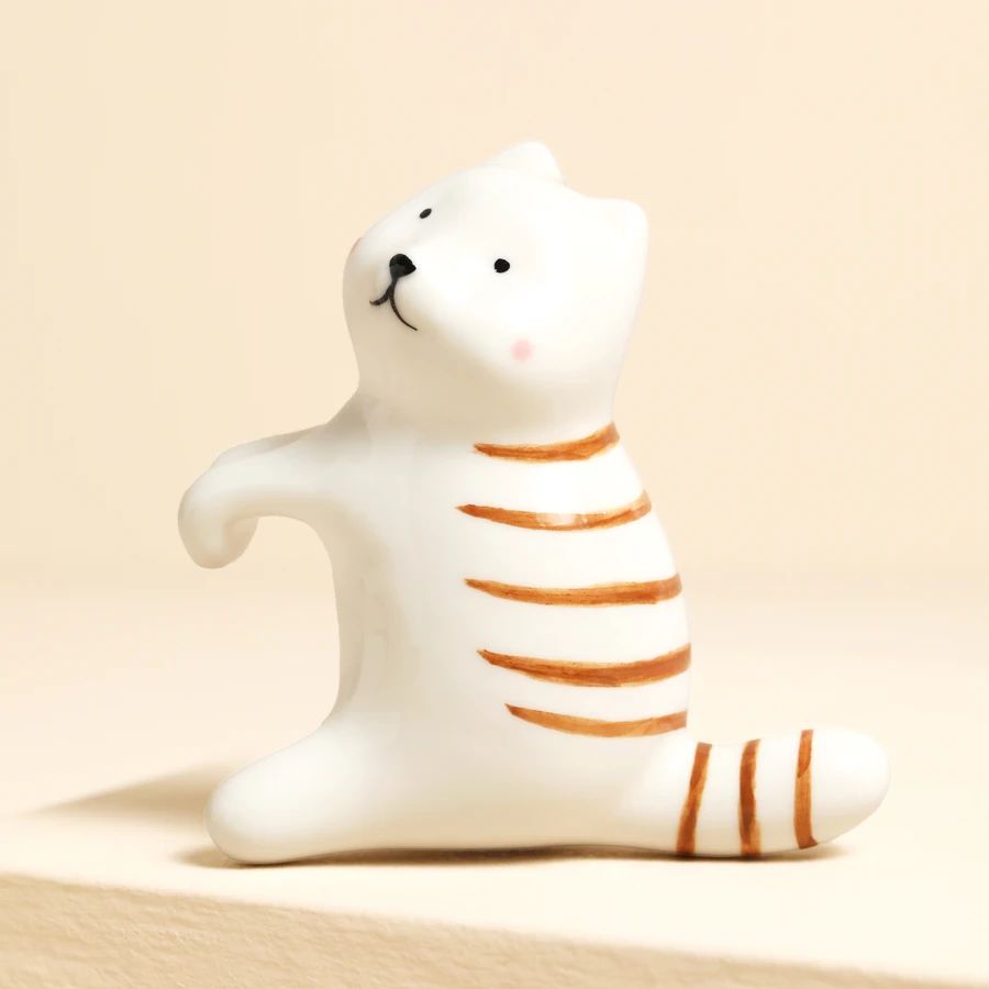 A small white cat figurine with orange stripes, sitting on a beige surface against a warm beige background.