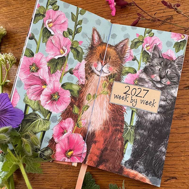 An open illustrated 2027 week by week planner. The cover art shows two cats among pink blossoms.