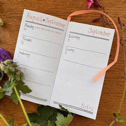 An open two-page weekly planner spread on a wooden surface, decorated with purple flowers and green leaves. 