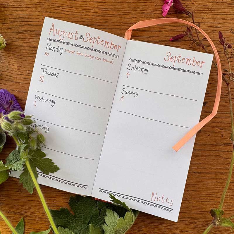 An open two-page weekly planner spread on a wooden surface, decorated with purple flowers and green leaves. 