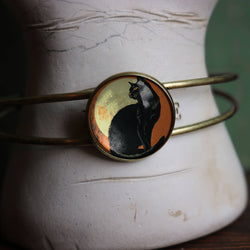 A brass bangle bracelet with a round cabochon featuring a black cat silhouette against a warm orange background