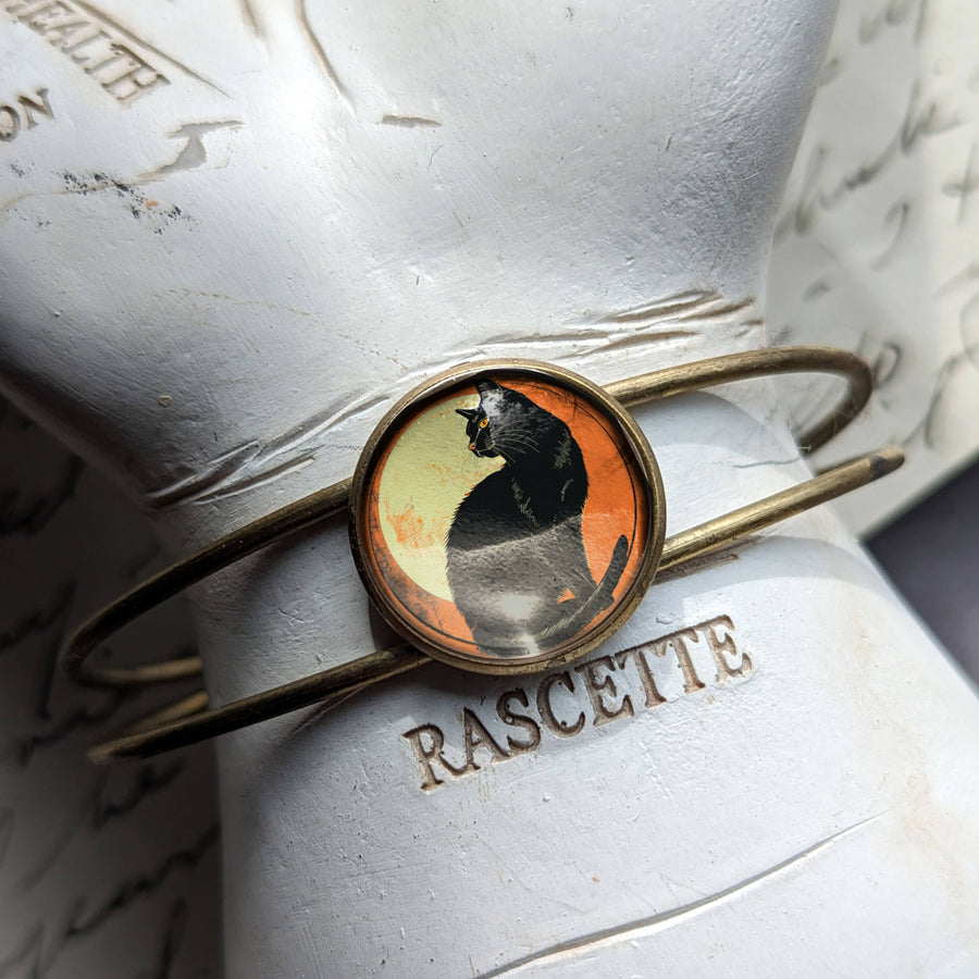 A thin gold-toned cuff bracelet with a round cabochon featuring a black cat silhouette against a warm orange background