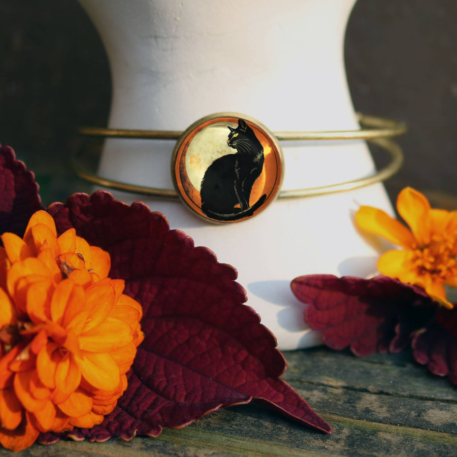 A gold bangle bracelet with a circular cabochon featuring a black cat silhouette against a warm yellow background