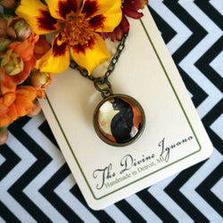 A round pendant necklace on a chain. The pendant shows a black cat silhouette against a warm orange-brown background.