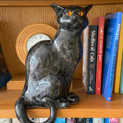 A glossy black ceramic cat figurine with orange eyes sits on a wooden bookshelf next to books and a clock