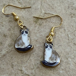 A pair of gold-tone dangle earrings featuring small cat-shaped charms with a white, grey, and black pattern