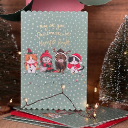 A festive Christmas greeting card featuring four illustrated cats wearing red holiday hats and scarves on a pale green polka-dot background with gold text reading 'May all your Christmas trees be Mums & Bright.'