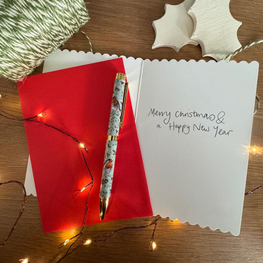 A decorative pen with a white and gray patterned barrel and gold accents rests on an open white card that reads 'Merry christmas & a Happy New Year',