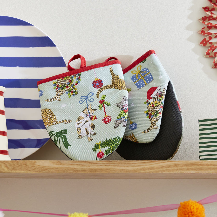 Pair of festive oven mitts with Christmas cat design on a wooden shelf.