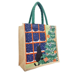 A jute tote bag with green rope handles, decorated with a Christmas window scene showing snow outside, a black cat silhouette, and a decorated Christmas tree 