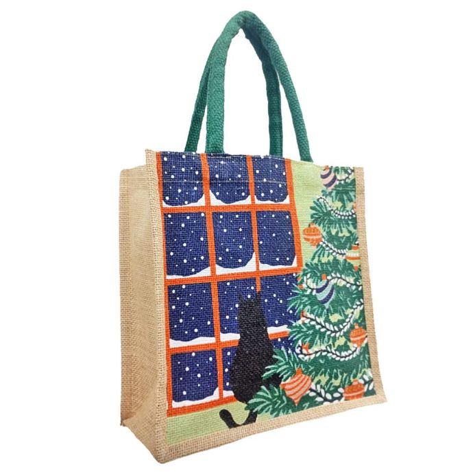 A jute tote bag with green rope handles, decorated with a Christmas window scene showing snow outside, a black cat silhouette, and a decorated Christmas tree 