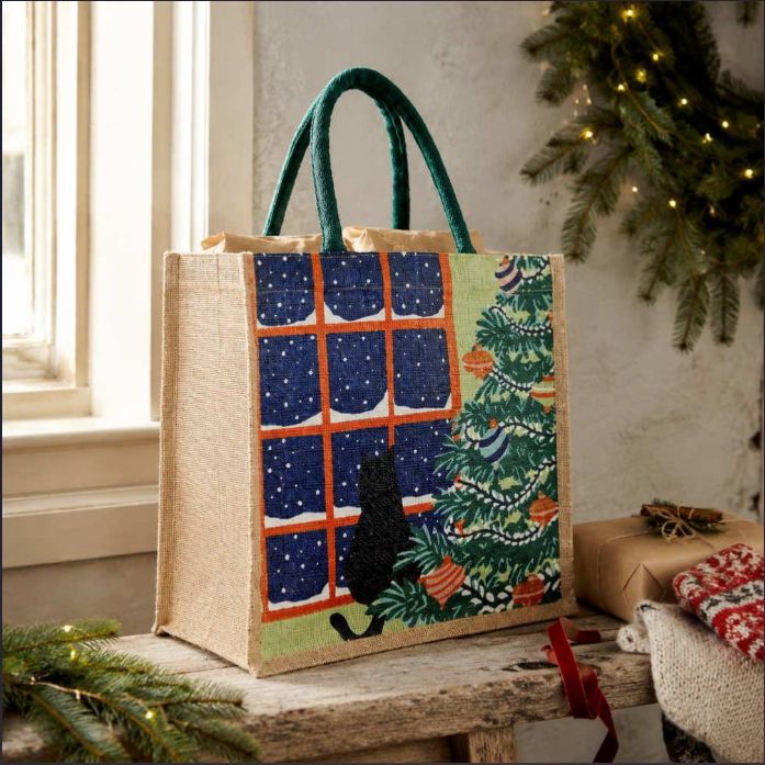 A jute tote bag with dark green handles sits on a rustic table. The bag features a printed design with a window scene showing a black cat, a snowy night sky, and a decorated Christmas tree.