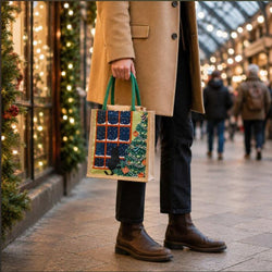 A person holds a Christmas-themed reusable shopping bag with green handles, featuring a black cat, festive window and tree illustration