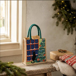 A jute gift bag with teal handles stands on a rustic wooden table. The bag features a printed design of a Christmas tree, a black cat by a window, and blue starry window panes.