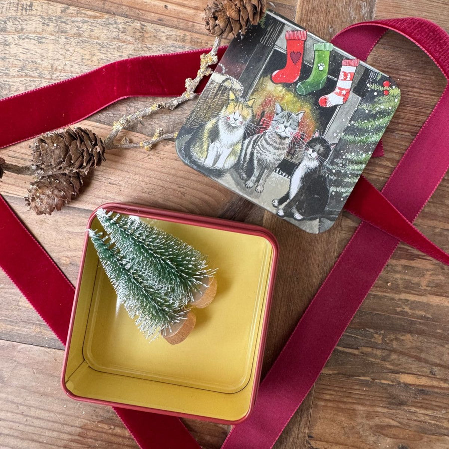 A square tin gift box with a festive Christmas design featuring cats and stockings, displayed on a rustic wooden table with a red velvet ribbon