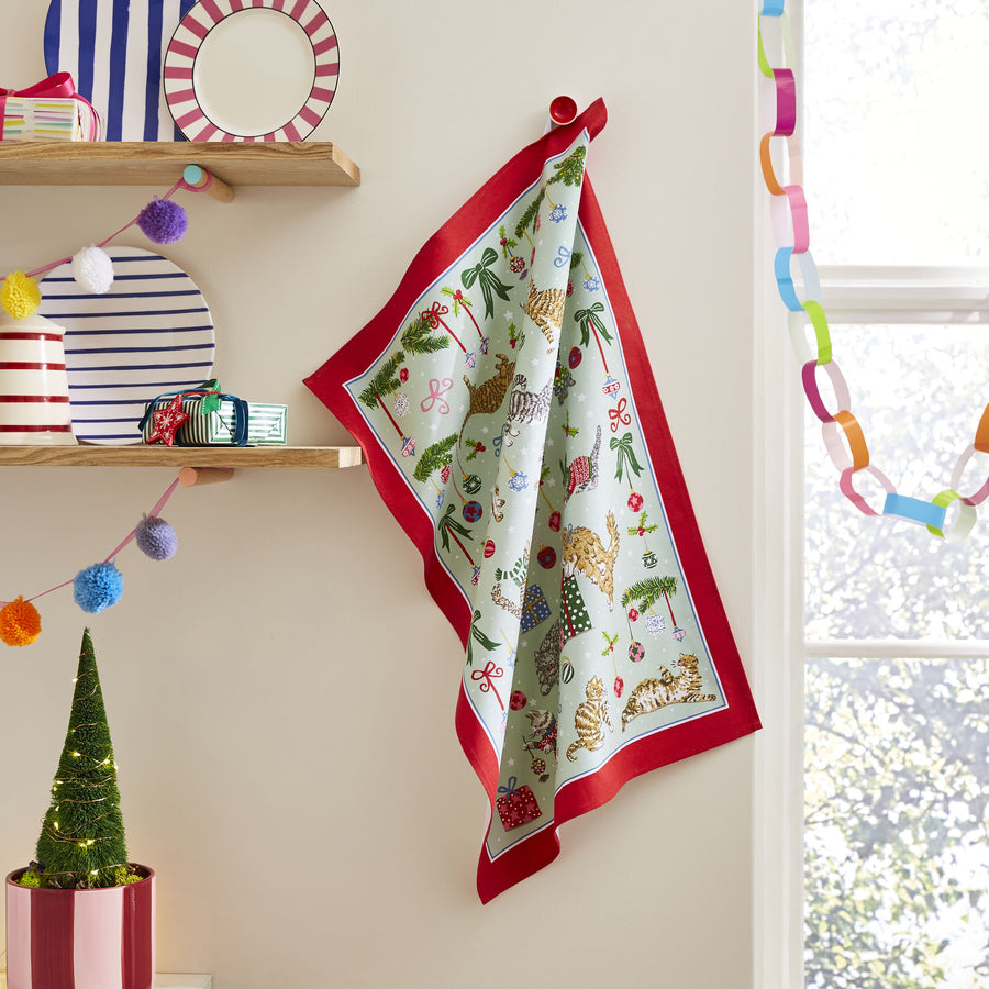 Decorative Christmas-themed  tea towel featuring cats and baubles with red border hanging on a wall.