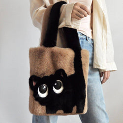 A person holds a plush faux-fur tote bag shaped like a black cat face, with tan side panels and black handles