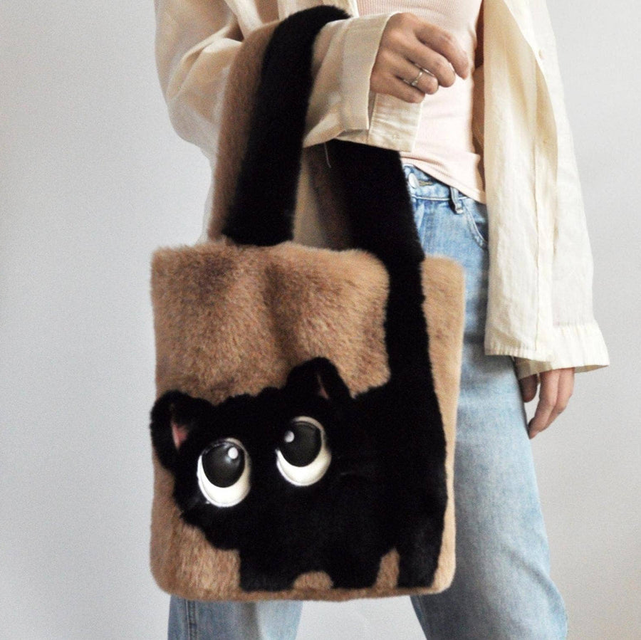 A person holds a plush faux-fur tote bag shaped like a black cat face, with tan side panels and black handles
