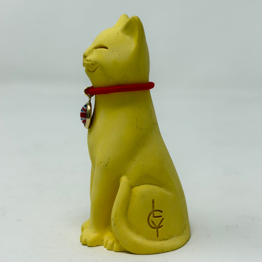 A smiling yellow lucky cat figurine wearing a red collar with a round Union Jack charm, photographed against a plain light gray background - side view with LCY logo visible