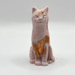 An aubergine coloured lucky cat figurine with rose gold leaf accents on its forehead, chest, and paws, standing against a neutral light grey background.