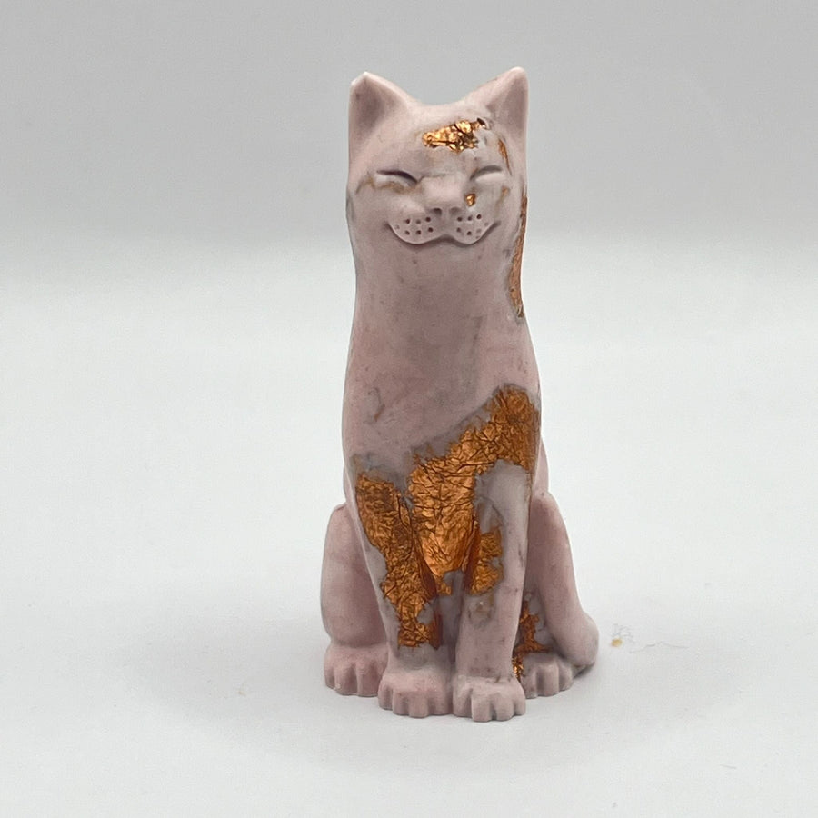 An aubergine coloured lucky cat figurine with rose gold leaf accents on its forehead, chest, and paws, standing against a neutral light grey background.