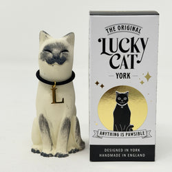 A  Lucky Cat figure hand painted to look like a blue point Siamese with a navy collar and L charm. Sitting up next to its retail packaging lablelled The Original Lucky Cat York.