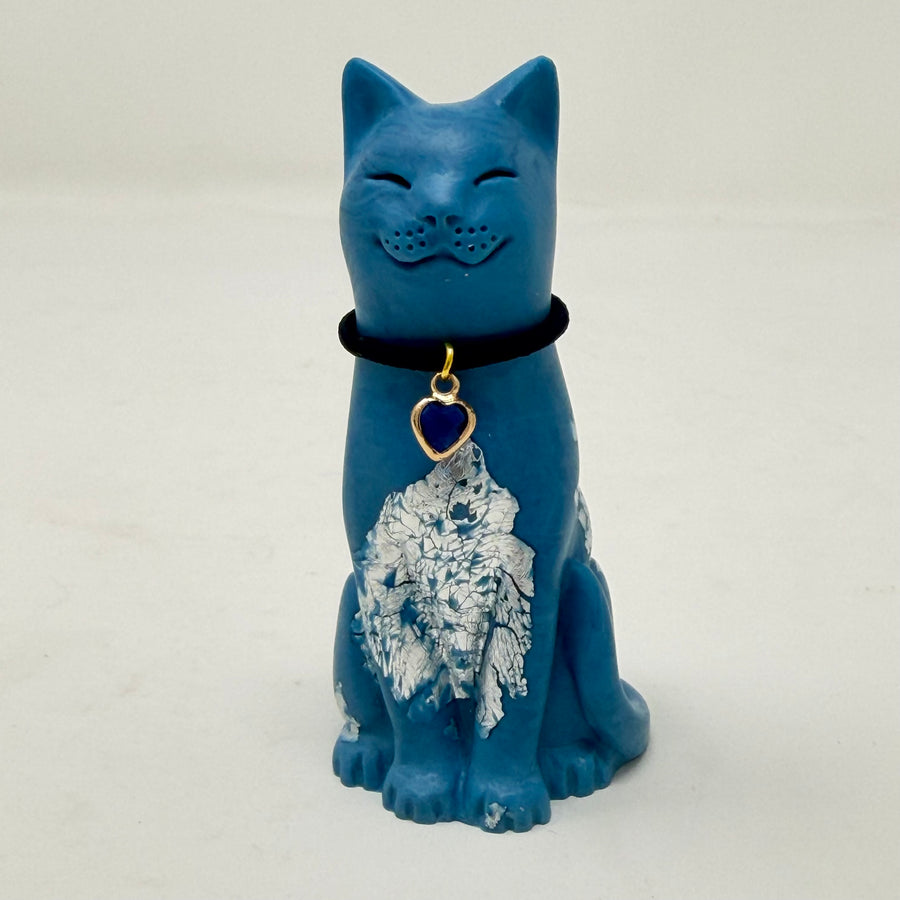 A blue smiling lucky cat figurine with silver foiled details and a black collar with blue heart charm on a light grey background.