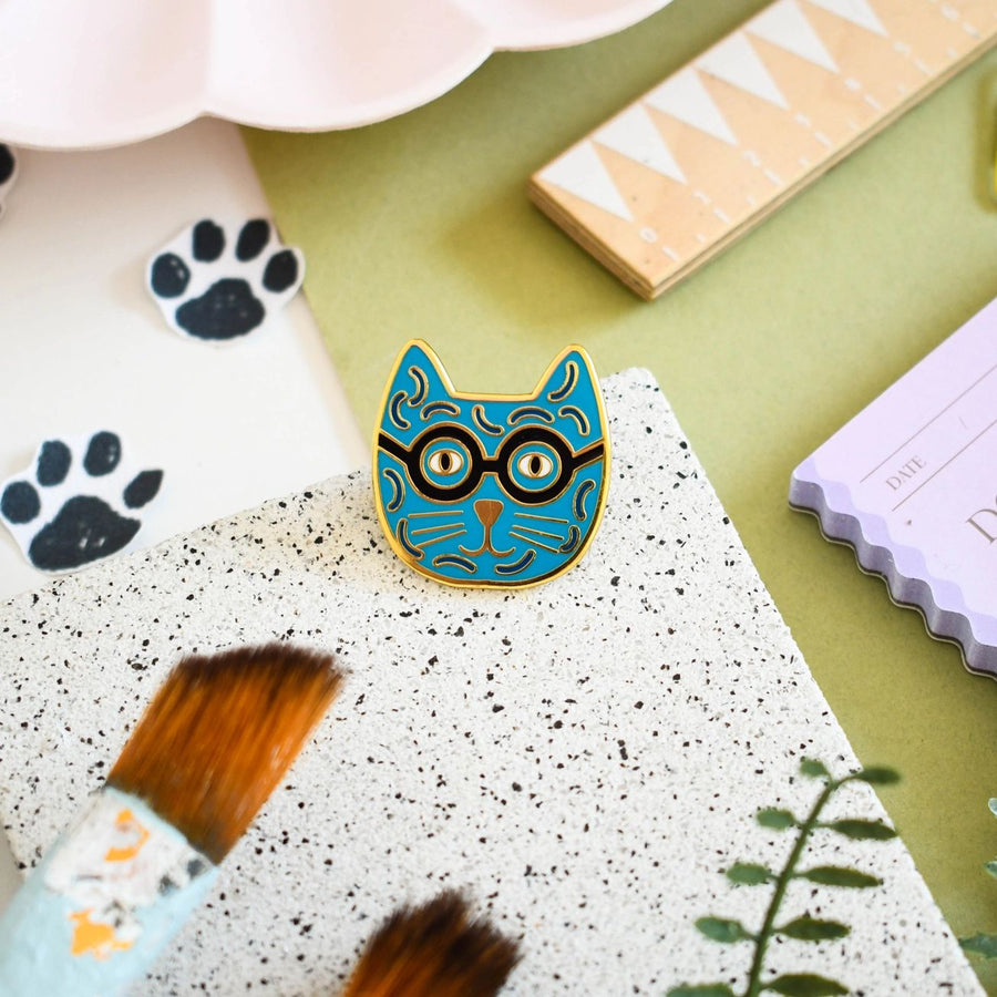 An enamel pin badge with a blue and yellow cat design wearing glasses