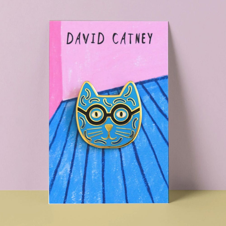 An enamel pin badge with a blue and yellow cat design wearing glasses on a illustrated backing card, labelled 'DAVID CATNEY'