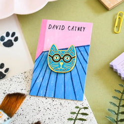 An enamel pin badge with a blue and yellow cat design on a illustrated backing card, labelled 'DAVID CATNEY'