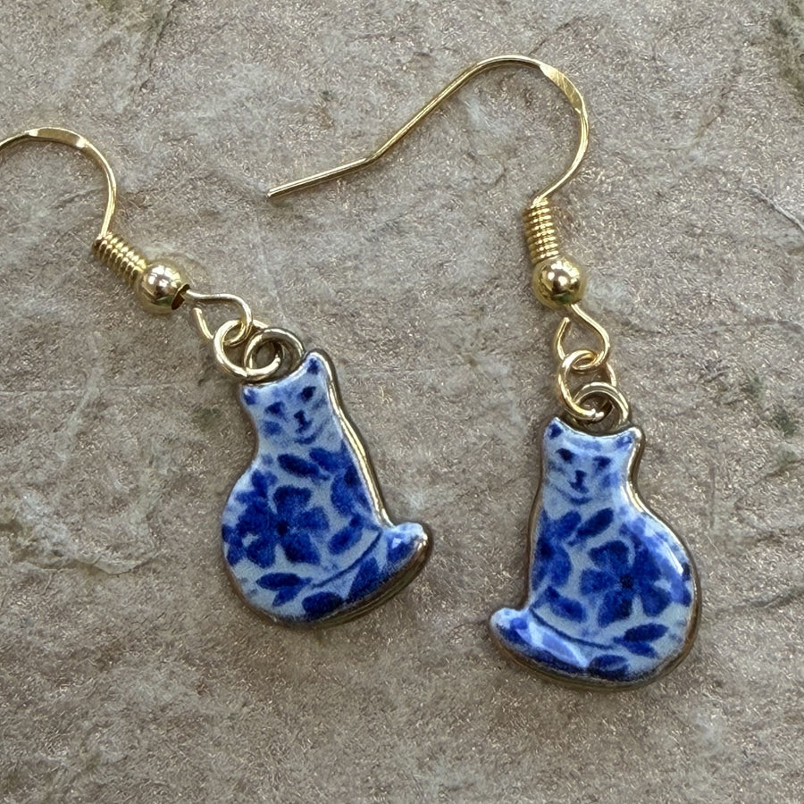 A pair of dangling earrings with blue and white patterned cat charms and gold-tone fishhook wires, displayed on a textured gray stone surface.