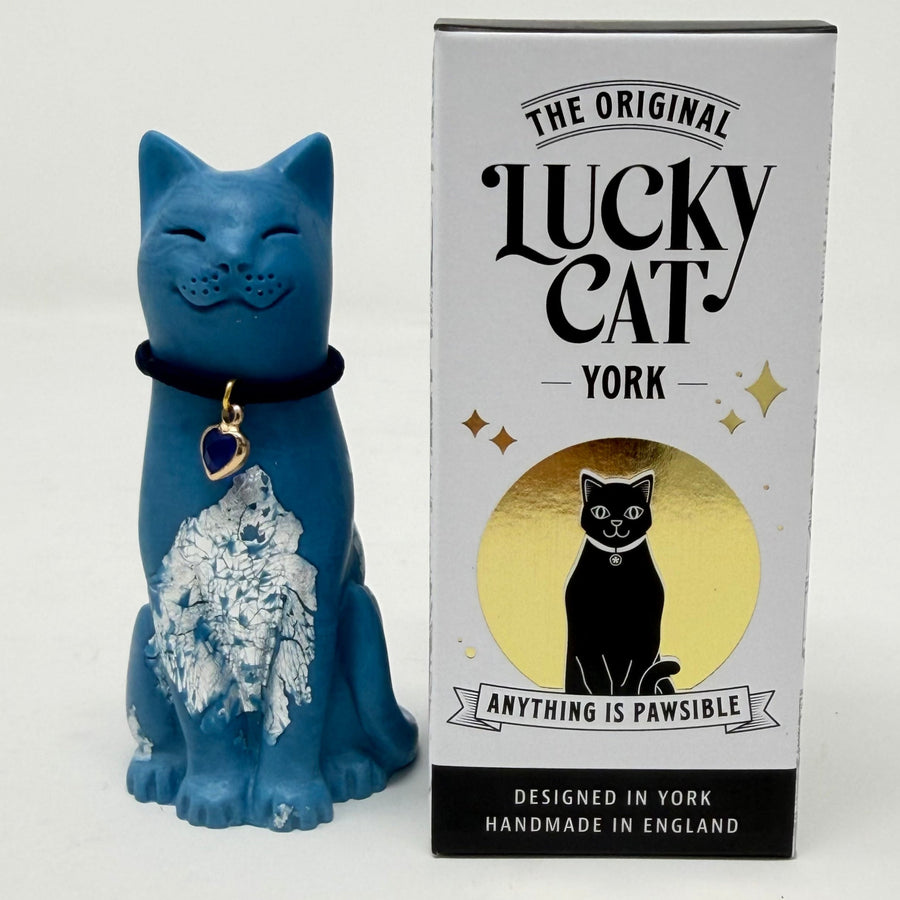 A blue cat figurine with silver foiled details and a black collar  with blue gemstone heart charm stands next to its white and black retail box labelled 'The Original Lucky Cat York'.