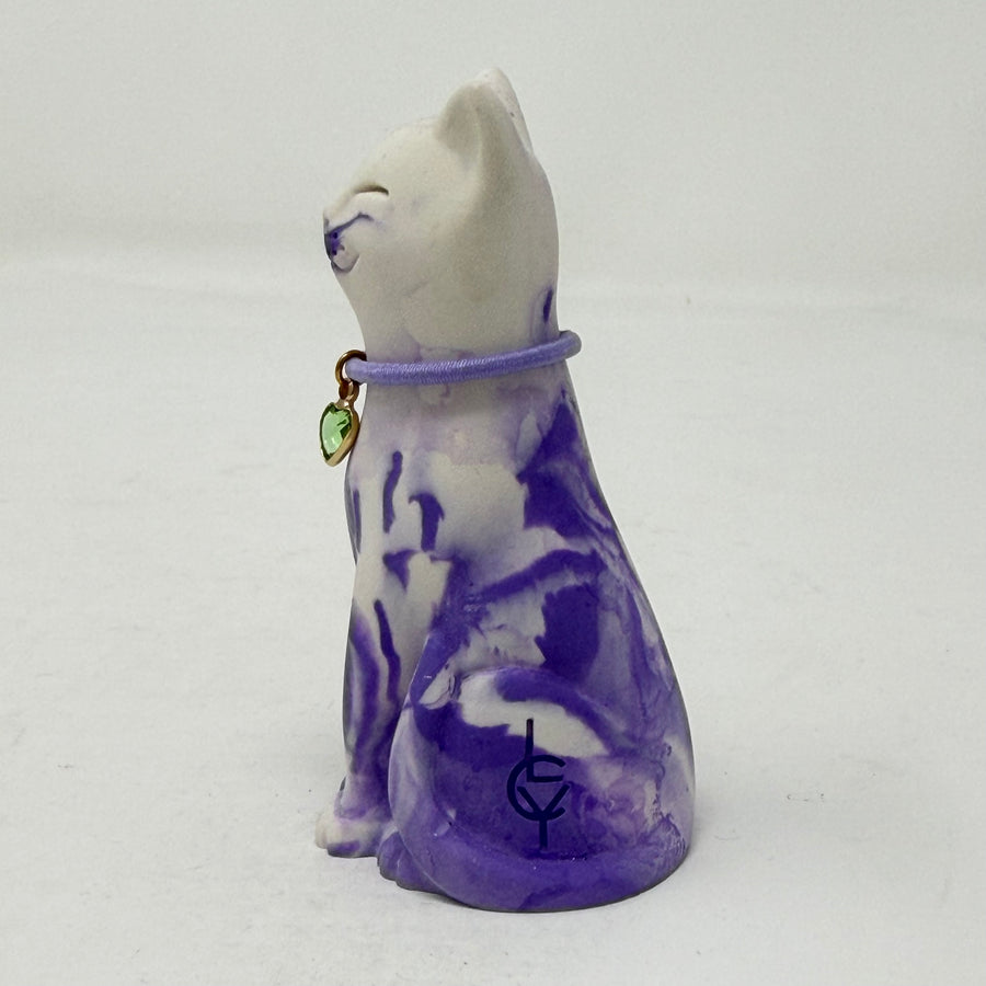 A lavender and white and marbled cat figurine wearing a purple collar with a green heart charm. Side view.