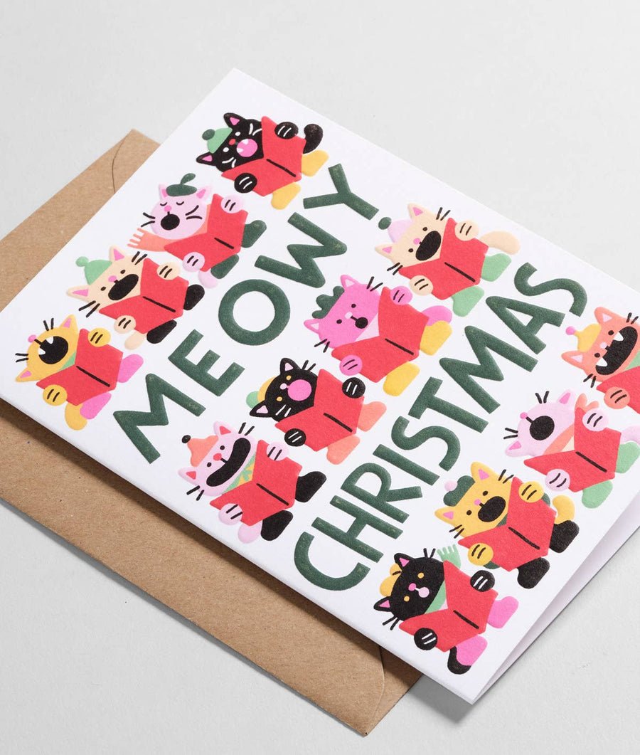 A white Christmas greeting card with the words 'MEOWY CHRISTMAS' in large dark green letters, surrounded by colorful cartoon cat characters in red, pink, yellow, and black clothing. 