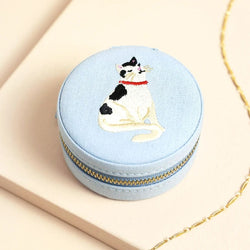 A round light blue fabric jewelry box with a gold zipper, featuring an embroidered black-and-white cat with a red collar on top