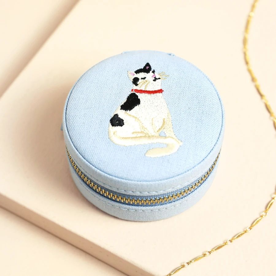 A round light blue fabric jewelry box with a gold zipper, featuring an embroidered black-and-white cat with a red collar on top
