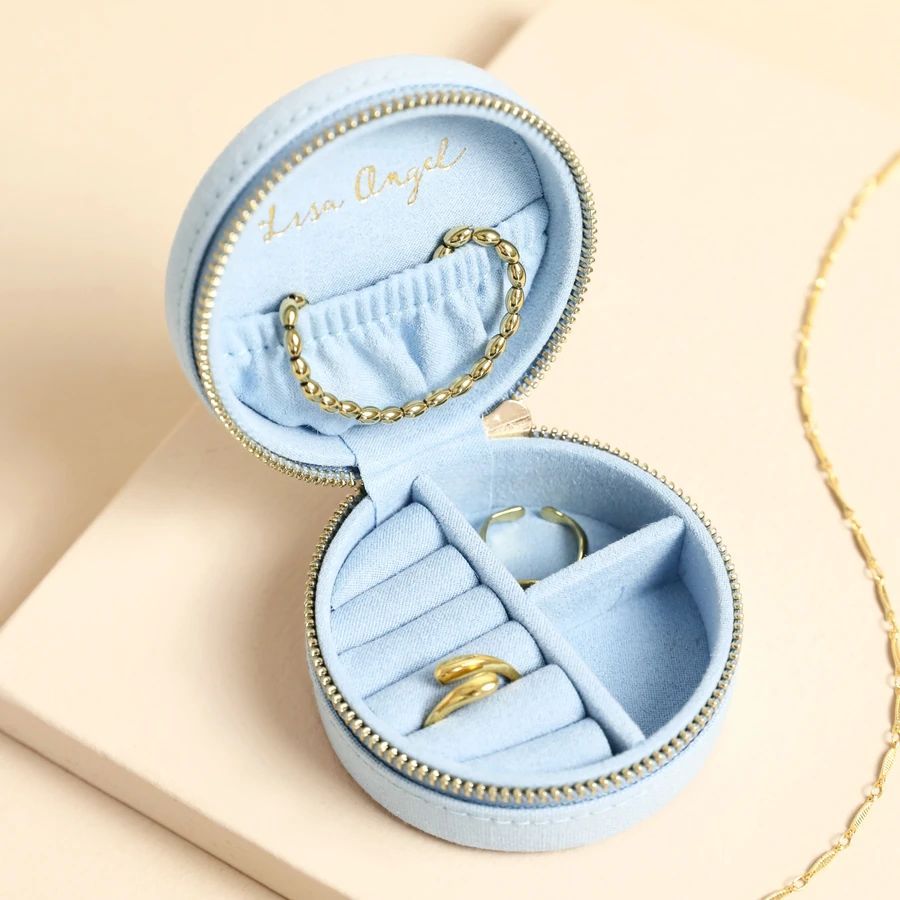 An open round light blue jewelry box with a zipper, holding gold rings and a gold chain necklace, photographed on a soft beige surface.
