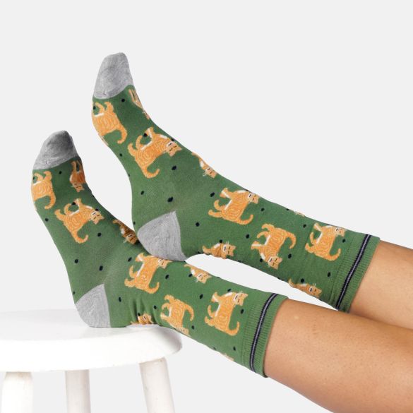 A pair of green crew socks with an orange tabby cat pattern and grey toes/heels, worn on feet resting on a stool