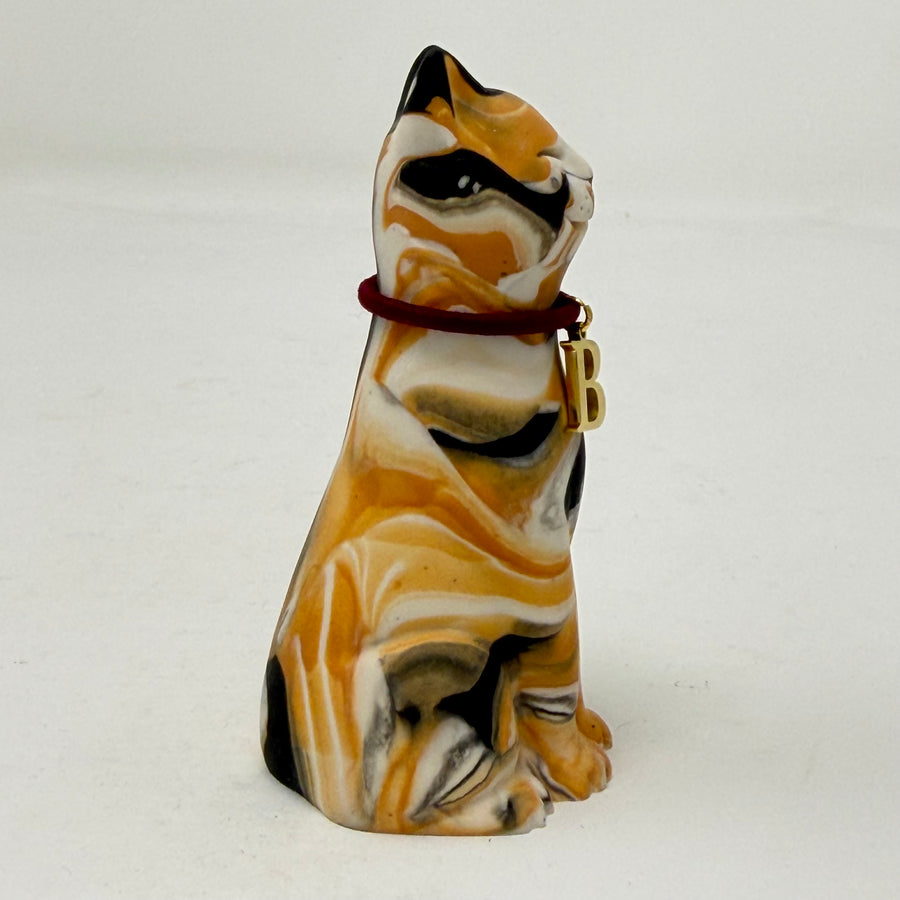 A marbled tortoiseshell Lucky Cat figurine wearing a burgundy collar with a gold letter B charm charm. Side view.