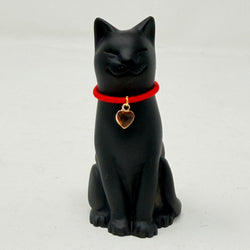 A black cat figurine wearing a bright red collar with a small gold heart-shaped charm, photographed against a plain light grey background.
