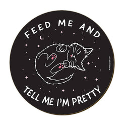 A round black coaster with a cute cartoon sleeping cat in the center, surrounded by white text that reads 'FEED ME AND TELL ME I'M PRETTY' 