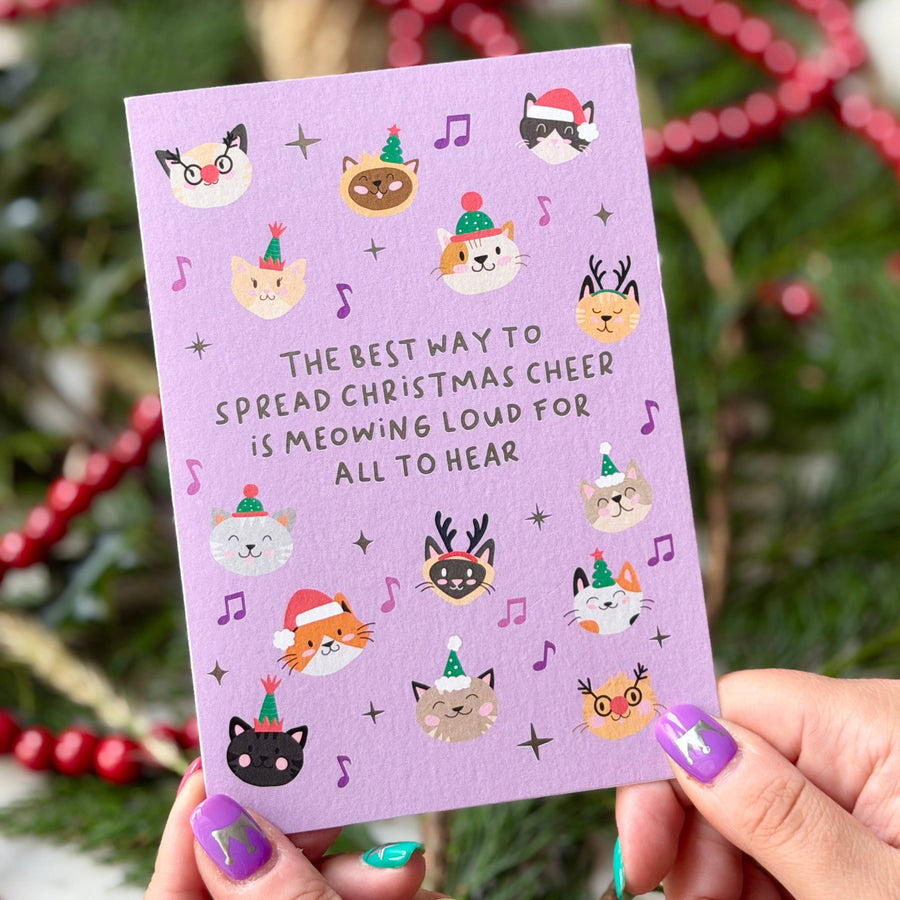A person holds a lavender Christmas greeting card with illustrations of cats in holiday hats and the text 'THE BEST WAY TO SPREAD CHRISTMAS CHEER IS MEOWING LOUD FOR ALL TO HEAR'. 