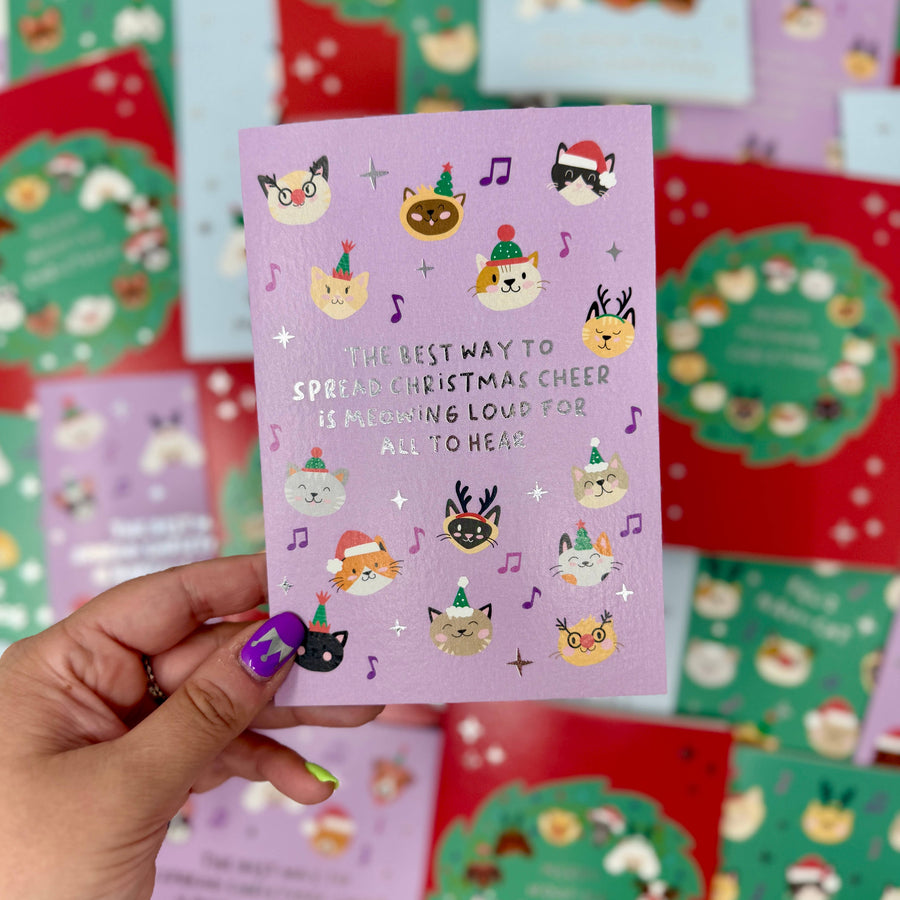 A hand with purple nail polish holds a lavender Christmas greeting card featuring cute illustrated cats in holiday hats and silver foil text. 