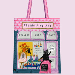 A tote bag featuring a whimsical illustration of a cat-themed art shop named 'Feline Fine Art', with pink patterned handles