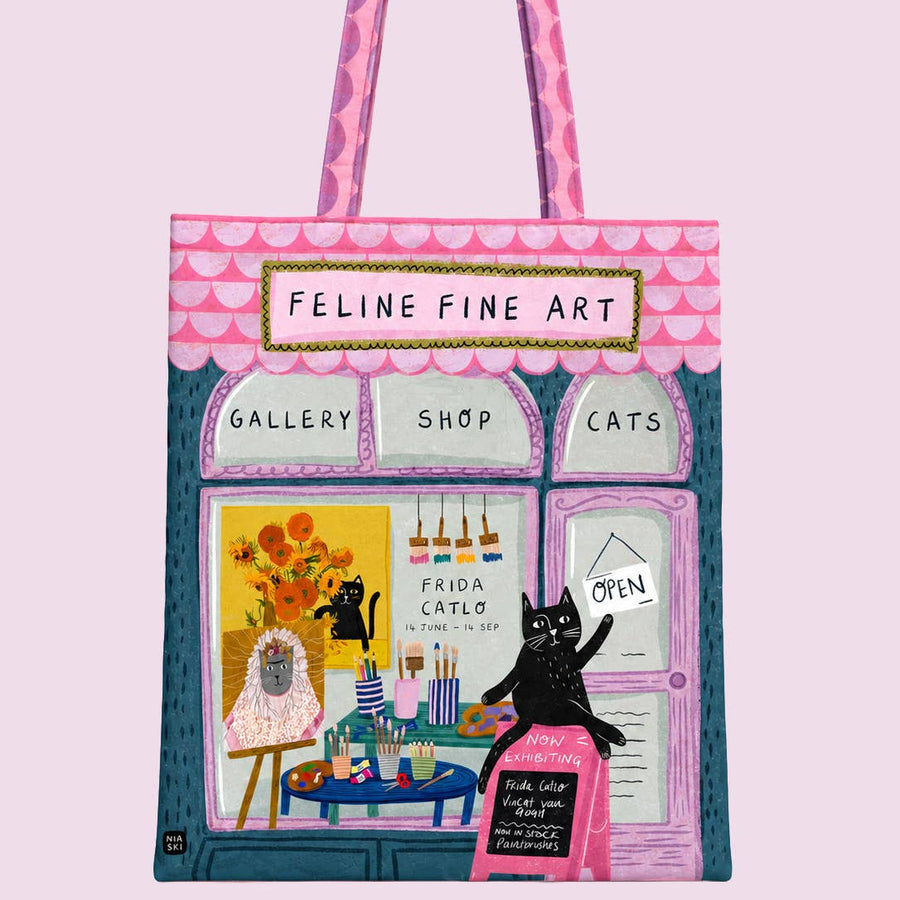 A tote bag featuring a whimsical illustration of a cat-themed art shop named 'Feline Fine Art', with pink patterned handles