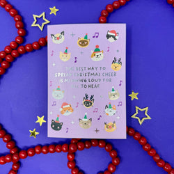 A lavender greeting card with cute illustrated cat faces and festive accents, placed on a purple background with red beaded garlands and gold star decorations.