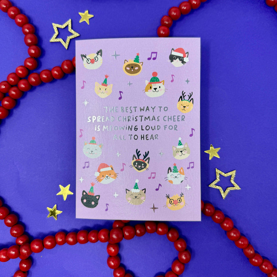 A lavender greeting card with cute illustrated cat faces and festive accents, placed on a purple background with red beaded garlands and gold star decorations.