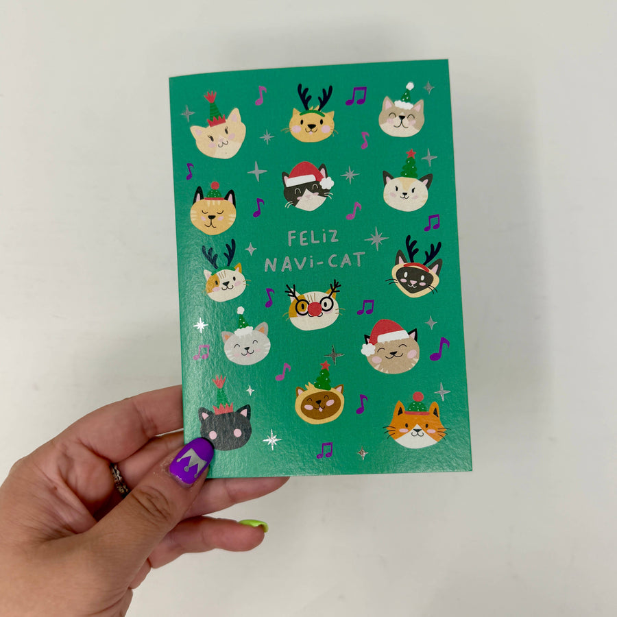 A hand holds a green Christmas-themed greeting card that reads 'FELIZ NAVI-CAT'. The card features cute cartoon cat faces wearing festive holiday accessories like Santa hats, reindeer antlers, and elf glasses