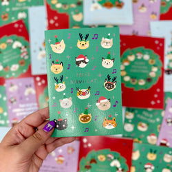 A hand holds a green greeting card featuring cute cat illustrations in holiday hats, with the text 'FELIZ NAVI-CAT'. 